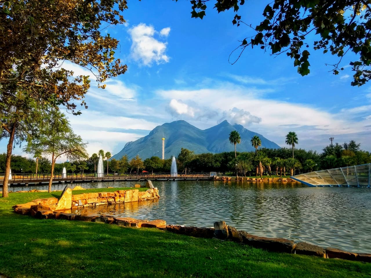 Paisaje natural que incluye un lago con palmeras alrededor, fuentes, y montañas al fondo bajo un cielo azul con algunas nubes. Perfecto para disfrutar de la tubería de Diámetros Mayores en entornos industriales en México.