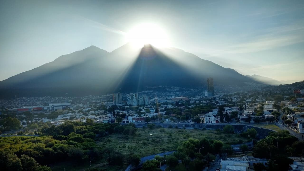 Vista panorámica de una ciudad con montañas al fondo, donde el sol se asoma detrás de las montañas, creando rayos de luz que se extienden sobre el paisaje urbano y natural, resaltando la vegetación y la infraestructura.