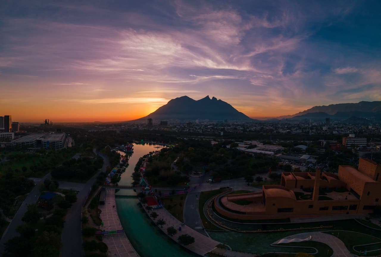 Atardecer en Monterrey, con montañas imponentes al fondo y un río que atraviesa un parque. Se pueden ver edificios y vegetación, mientras el cielo presenta tonos anaranjados y violetas. Destacan estructuras modernas en el primer plano.