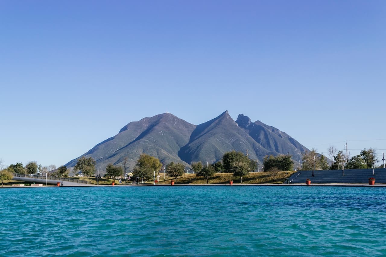 Vista de una imponente montaña con forma triangular, rodeada de un lago de aguas azuladas y un cielo despejado. En la base, se observan áreas verdes y árboles. La escena evoca tranquilidad y belleza natural, ideal para actividades al aire libre.