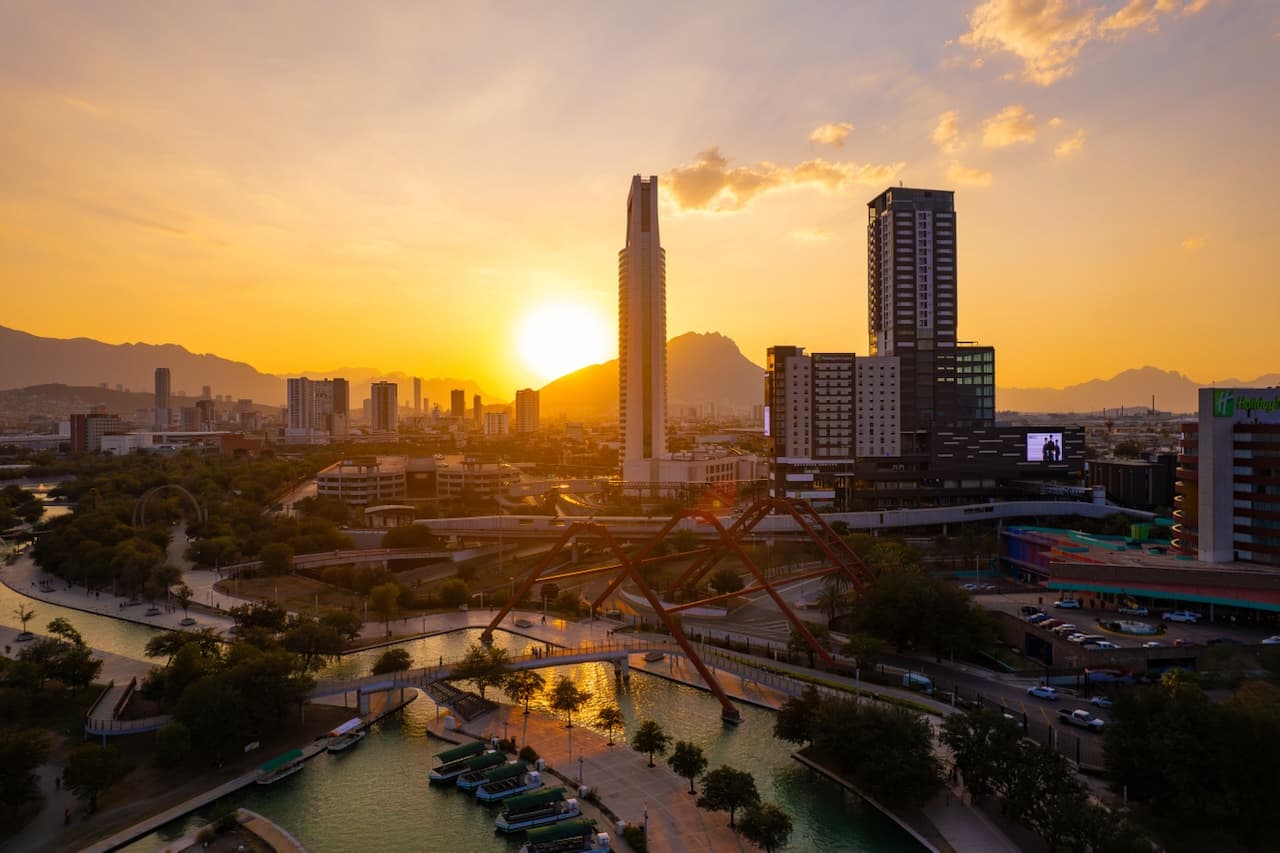 Vista panorámica de una ciudad al atardecer, con edificios modernos y un río en primer plano, rodeado de vegetación y montañas al fondo. Se destaca una estructura arquitectónica de diseño especial sobre el agua, mientras el sol se oculta en el horizonte, creando un ambiente cálido y vibrante.