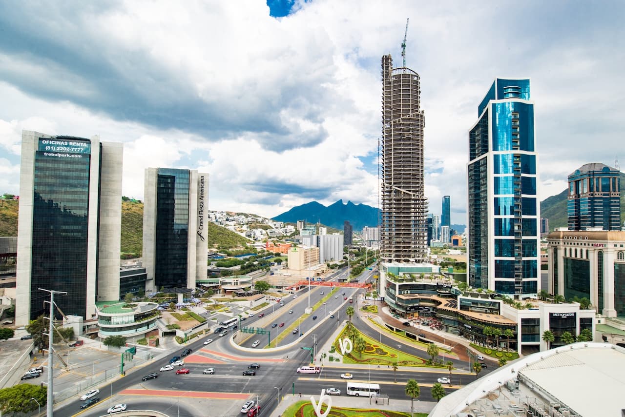 Vista panorámica de un área urbana moderna con varios edificios de gran altura, incluida una construcción en proceso, rodeada de montañas y con un tráfico vehicular visible. Se observan zonas verdes y espacios públicos en la intersección de importantes vías.