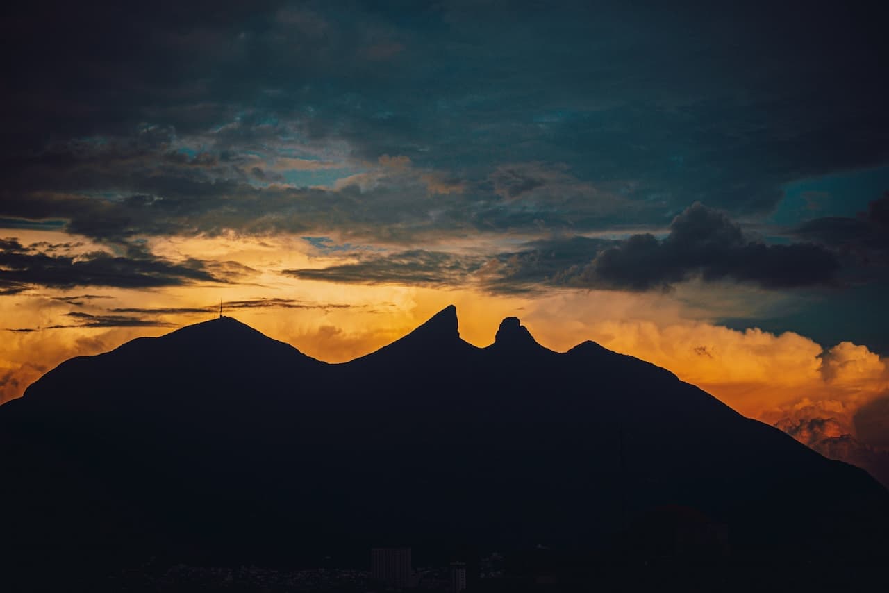 El paisaje presenta una silueta de montañas con picos distintivos, contrastando con un cielo lleno de nubes y un atardecer de colores cálidos, en un entorno natural que evoca tranquilidad y majestad.