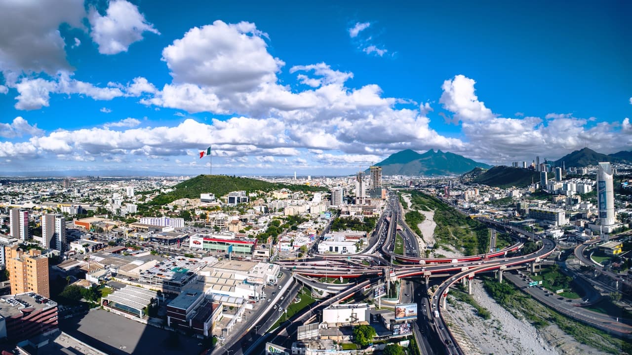 Vista panorámica de una ciudad con edificios, una montaña prominente y un gran puente en una intersección, destacando una bandera mexicana en un cerro. El cielo está parcialmente nublado y se observan diversas carreteras y tráfico. Se sugiere el uso de Tubería de Diámetros Mayores para las infraestructuras urbanas.