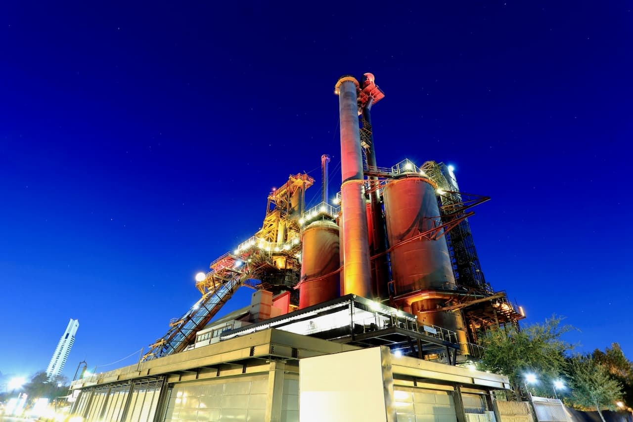 Planta industrial iluminada durante la noche, con chimeneas y tanques grandes, destacando su estructura metálica y el cielo estrellado de fondo, que sugiere su uso en procesos industriales complejos.