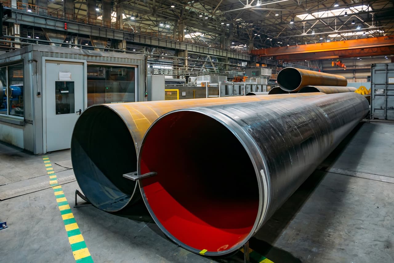 Se observa Tubería de Diámetros Mayores apiladas en un ambiente industrial, con una estructura metálica de fondo y una caja de control. Algunas tuberías tienen un acabado negro, mientras que otras presentan un interior rojo. El espacio está bien iluminado, lo que resalta las características de la tubería.